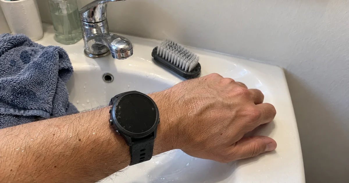 Garmin Forerunner s černým silikonovým řemínkem, zápěstí nad umyvadlem s kartáčkem a mýdlovou vodou