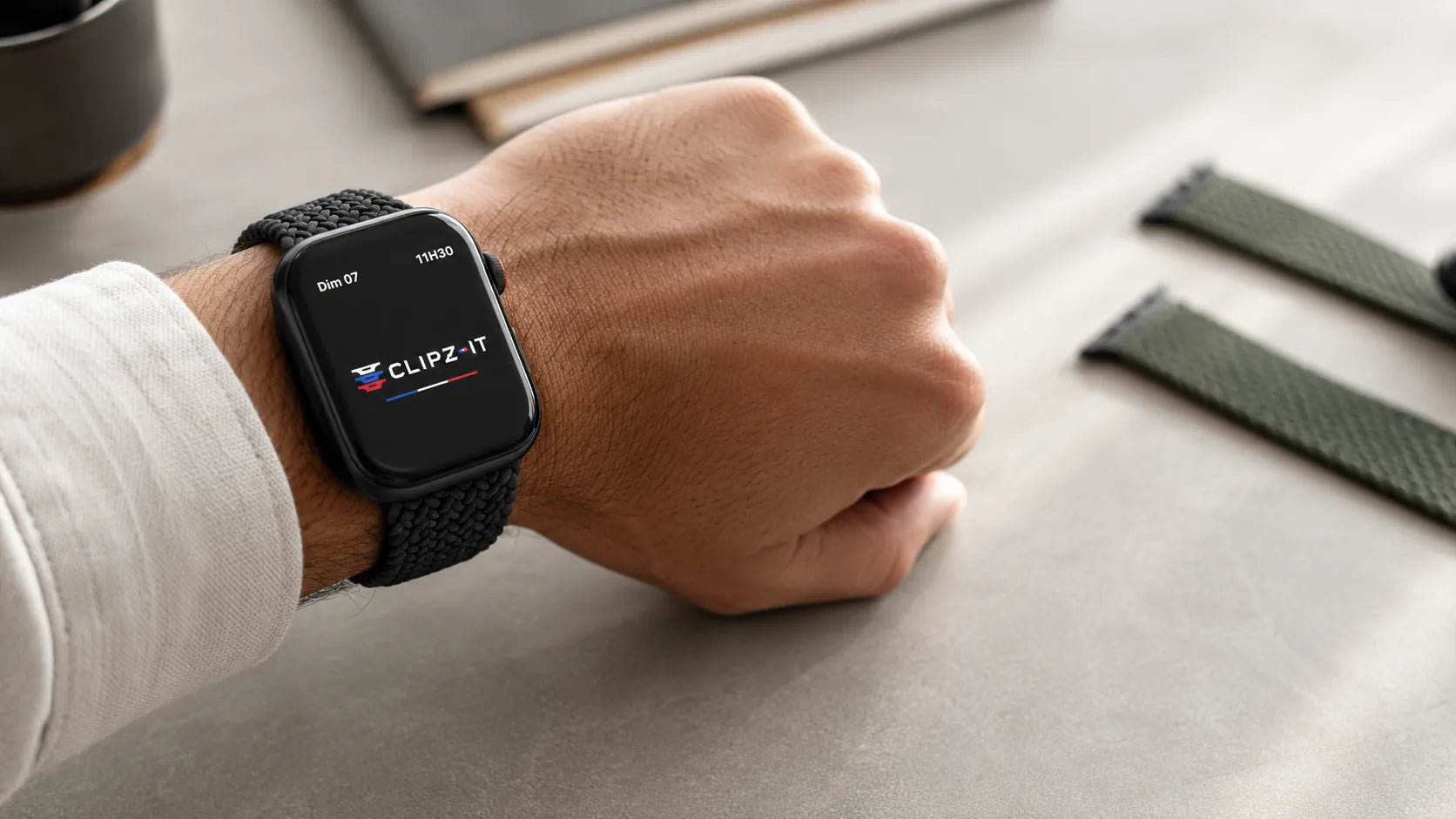 Poignet avec Apple Watch Series 10 et bracelet nylon tressé noir sur un bureau, un second bracelet posé à côté - Clipz-it Image